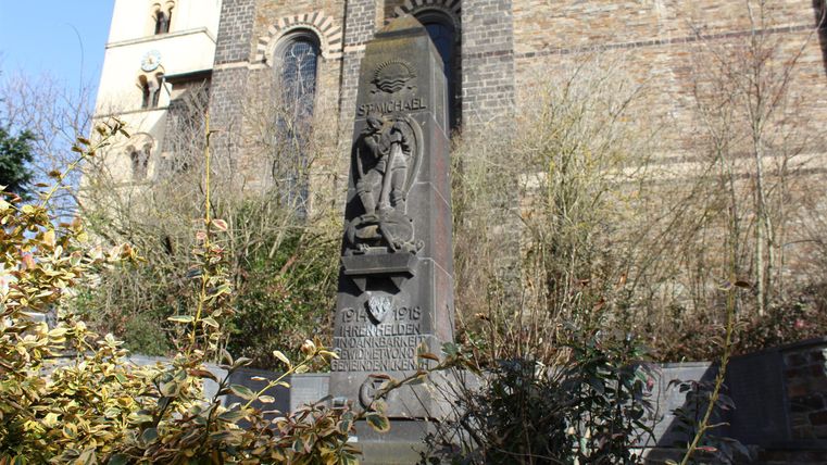 A statue stands in front of an old church, surrounded by bushes and plants. The architecture of the church is well-preserved and shows details in stone.