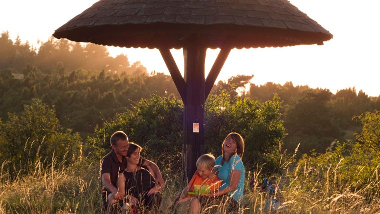 Familie sitzt unter einem hölzernen Unterstand in einer Wiese bei Sonnenuntergang.