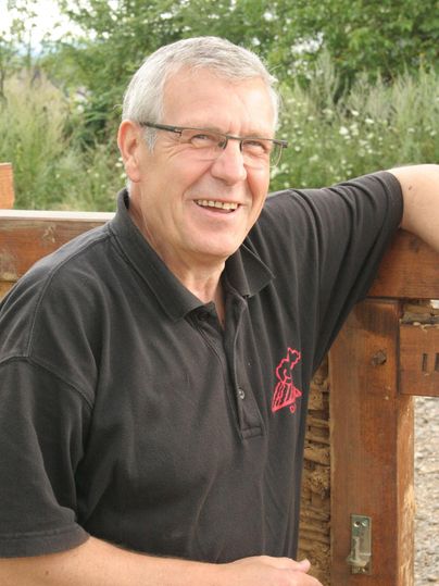 A smiling man stands by a wooden fence wearing a black shirt. In the background, green plants and trees are visible.