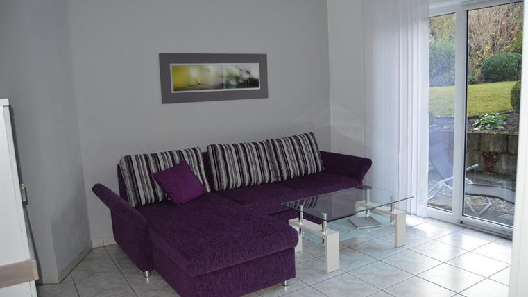 Ein modern eingerichtetes Wohnzimmer mit einem lila Sofa und einem Glastisch. Große Fenster lassen viel Tageslicht herein und bieten einen Blick ins Freie.