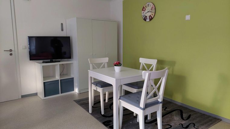 A bright room with a white table and four chairs. A television is mounted on the wall and there is a green accent area.