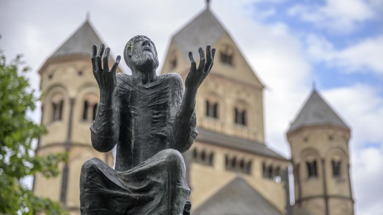 Eine Statue eines betenden Mönchs steht im Vordergrund. Im Hintergrund sind alte Gebäude mit Türmen zu sehen.