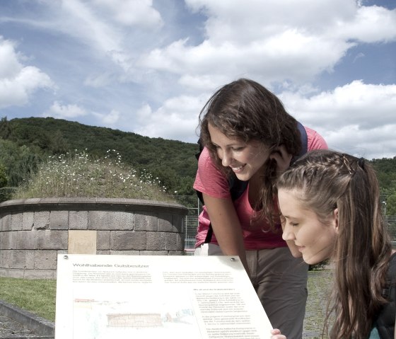 Zwei Frauen betrachten eine Informationstafel im Freien. Im Hintergrund ist ein rundes Steindenkmal und bewaldete H&uuml;gel zu sehen., &copy; Traumpfade/Kappest