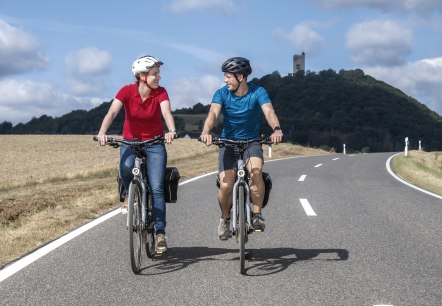 Zwei Fahrradfahrer fahren eine Stra&szlig;e entlang. Im Hintergrund die Burg Olbr&uuml;ck., &copy; Vulkanregion Laacher See, Klaus-Peter Kappest