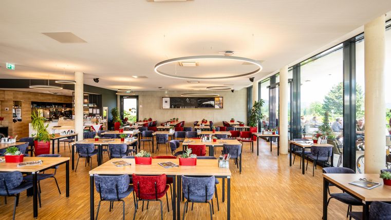 Ein modernes Restaurant mit hellem, einladendem Ambiente. Die Tische sind stilvoll gedeckt und umgeben von großen Fenstern mit Blick ins Freie.