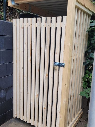 Eine Holzlagerbox mit vertikalen Latten und einem schrägen Dach. Die Box ist verschlossen und steht an einer Wand im Freien.