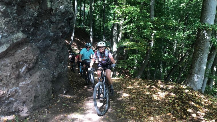 Eine Gruppe von Radfahrern fährt auf einem schmalen Pfad durch einen dichten Wald. Der Weg verläuft neben einem großen Felsen und ist mit Laub bedeckt.