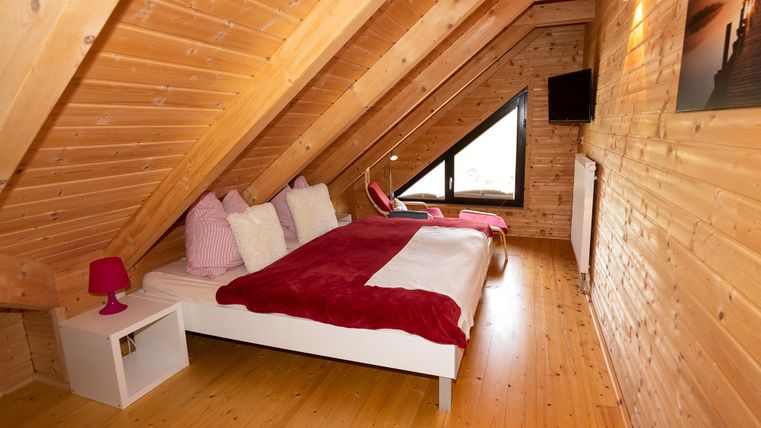 A cozy bedroom in the attic with wooden paneling. The room features a large bed, a television, and a window looking outside.