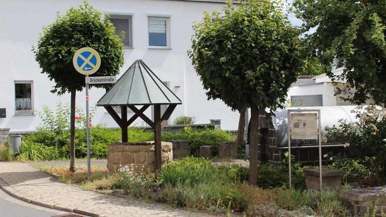 Ein schöner Brunnen mit einem Dach in einer kleinen Gemeinde. Im Hintergrund stehen einige grüne Bäume und ein Verkehrsschild.