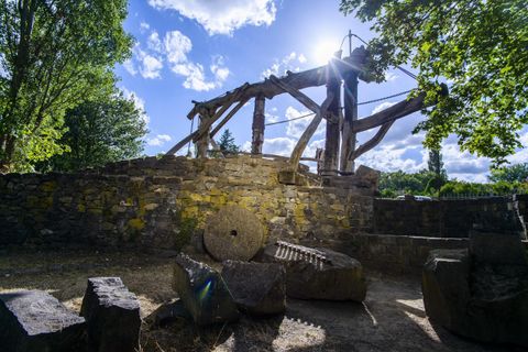 Eine alte Mühle steht inmitten von Bäumen, umgeben von verschiedenen Steinen. Die Sonne scheint durch die Wolken und beleuchtet die Szene.