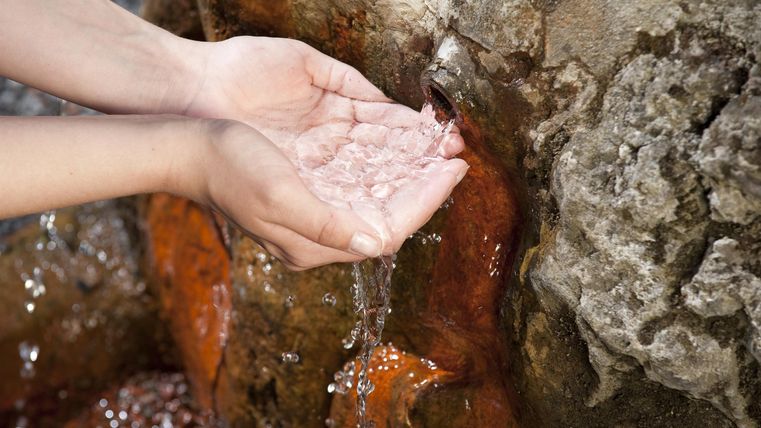 Hände fangen frisches Wasser aus einer Quelle. Im Hintergrund sind steinerne Felsen sichtbar.