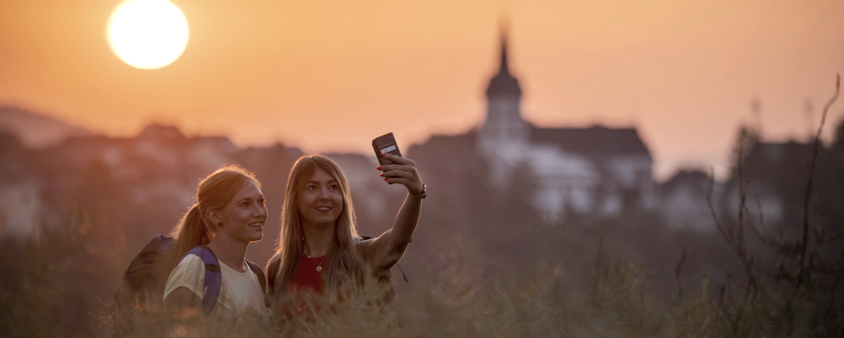 Twee mensen nemen een selfie in het groen voor een zonsondergang. Er staat een kerk op de achtergrond., &copy; Kappest_REMET