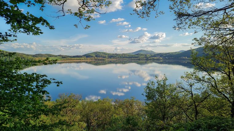Ein ruhiger See, umgeben von Bäumen und Hügeln, spiegelt den klaren Himmel und die Wolken wider. Die Natur wirkt friedlich und einladend.