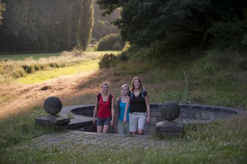 Three women are standing by a stone fountain in a green landscape. The surroundings are peaceful and surrounded by trees.