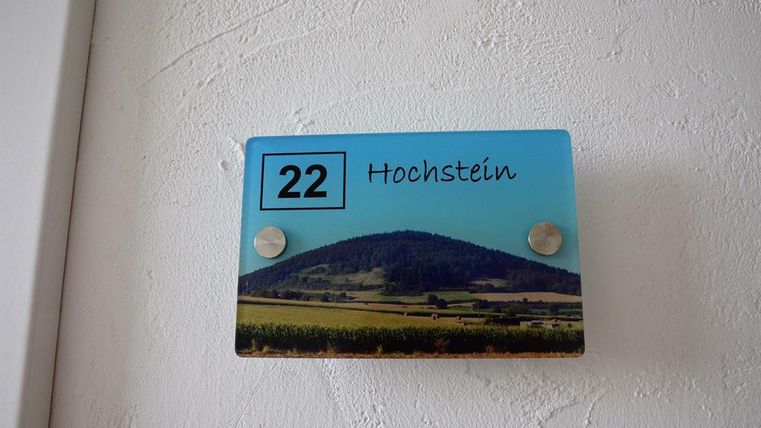 Ein Namensschild mit der Nummer 22 und dem Wort "Hochstein". Im Hintergrund ist eine Landschaft mit einem Hügel und Feldern dargestellt.