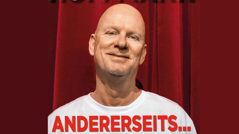 Ein lächelnder Mann steht vor einem roten Vorhang. Er trägt ein weißes T-Shirt mit einem auffälligen Schriftzug.