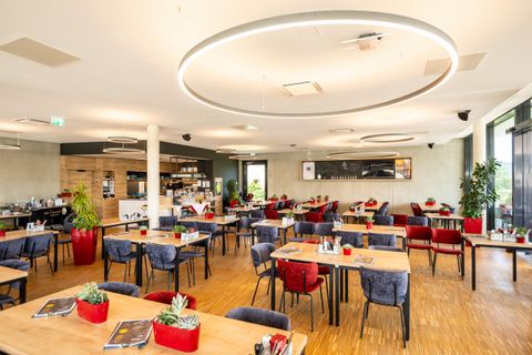 Ein modernes Restaurant mit vielen Tischen und Stühlen in verschiedenen Farben. Die Atmosphäre ist freundlich und einladend, mit Pflanzenakzenten und hellen Lichtquellen.