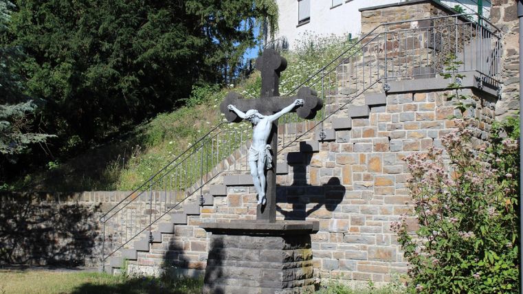A crucifixion sculpture stands in front of a stone staircase. In the background, trees and a building are visible.