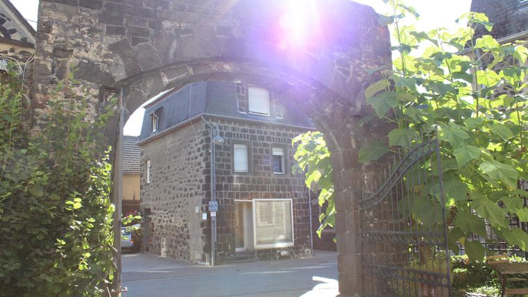 Ein schöner Steinbogen, der den Blick auf ein charmantes Haus in der Sonne freigibt. Im Vordergrund sind einige grüne Pflanzen zu sehen.