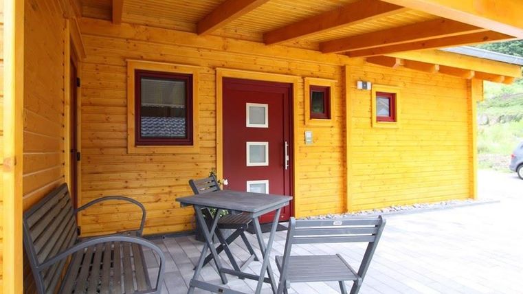 Eine gemütliche Holzterrasse mit einem Tisch und Stühlen. Die Fassade des Gebäudes ist aus hellem Holz mit einer roten Tür.