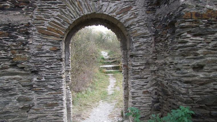 An old stone archway leading to a narrow path. In the background, green plants and a natural setting are visible.