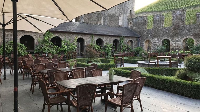Ein gemütlicher Innenhof mit Tischen und Stühlen unter Sonnenschirmen. Umgeben von Pflanzen und historischen Mauern, lädt der Platz zum Entspannen ein.