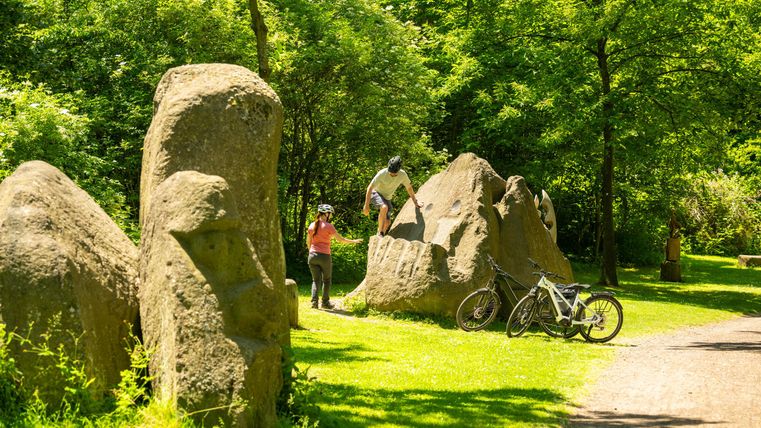 Eine grüne Parklandschaft mit großen Steinen und Radfahrern. Zwei Personen genießen die Natur und eine aktive Zeit im Freien.