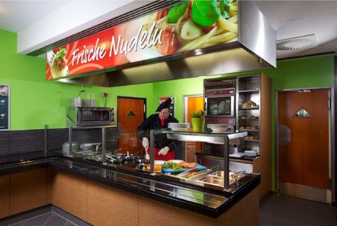 Eine moderne Küche mit einem Koch, der frische Nudeln zubereitet. Im Hintergrund sind grüne Wände und ein dekoratives Schild zu sehen.