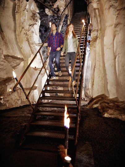 Ein Paar läuft die Treppe in einer Höhle hinunter. Fackeln beleuchten die steinernen Wände.
