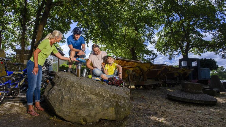 A family is sitting on a large rock and enjoying their time outdoors. In the background, bicycles and green spaces can be seen.
