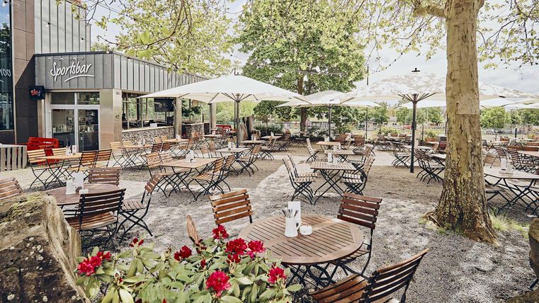 Ein einladendes Café mit vielen Tischen und Sonnenschirmen im Freien. Umgeben von Pflanzen und einem schönen grünen Bereich.