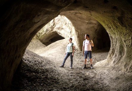 Zwei Wanderer stehen in einer gro&szlig;en H&ouml;hle mit erdigen W&auml;nden und &Ouml;ffnungen, die von nat&uuml;rlichem Licht erhellt werden., &copy; Eifel Tourismus GmbH, Dominik Ketz