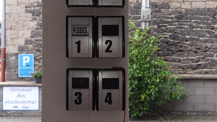 Ein schwarzes Informationssystem mit mehreren Schaltern ist an einer Wand montiert. Im Hintergrund sieht man grüne Pflanzen und eine Steinmauer.