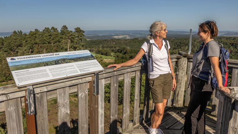 Zwei Wanderer stehen auf einer Aussichtsplattform und genießen die Aussicht auf die Landschaft. Im Hintergrund ist ein Schild mit Informationen über die Umgebung zu sehen.