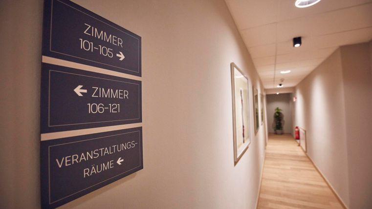 Ein heller Flur mit Beschilderungen zu verschiedenen Zimmern und Veranstaltungsräumen. Der Boden ist aus Holz und die Wände sind neutral gestrichen.