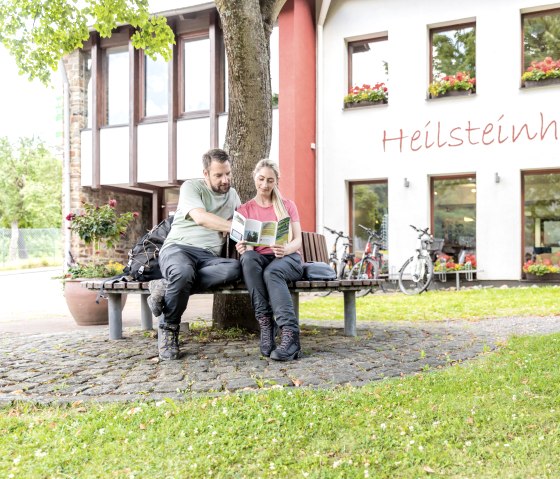 Twee mensen zitten op een bankje voor het Heilsteinhaus in Einruhr en lezen een brochure. Er staan fietsen geparkeerd voor het gebouw, dat is versierd met bloemen., &copy; -@Eifel Tourismus GmbH, AR-shapefruit-AG