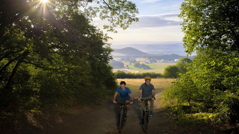 Two cyclists are enjoying a picturesque path in nature. Which is surrounded by trees and offers a beautiful view of the valley.
