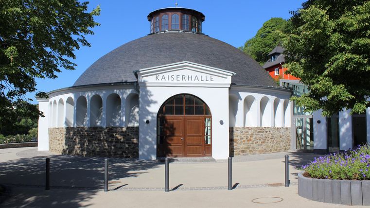 A historical round hall with a covered roof and large windows. Surrounded by trees and a clear blue sky.