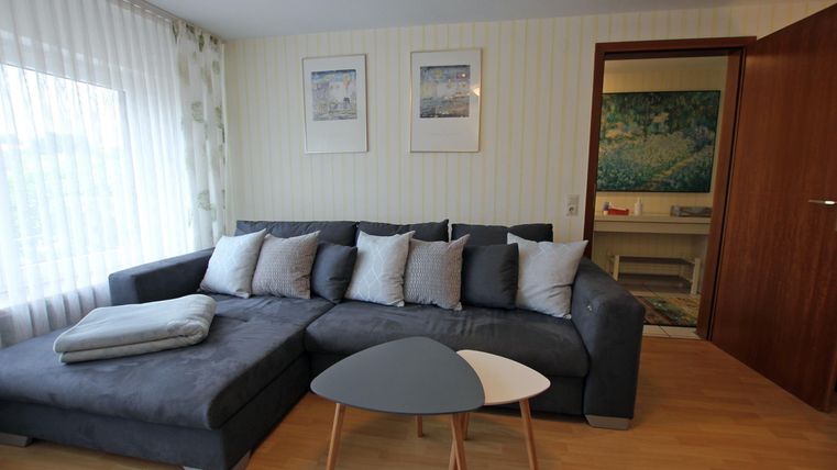 Ein gemütliches Wohnzimmer mit einem grauen Sofa und vielen Kissen. Im Hintergrund sind Bilder an der Wand und ein Zugang zu einem weiteren Raum sichtbar.
