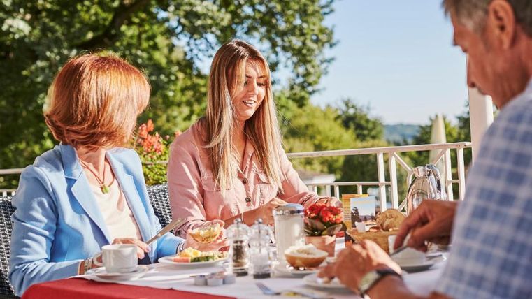 Eine Gruppe von drei Personen sitzt an einem Tisch im Freien und genießt ein Frühstück. Die Atmosphäre ist freundlich und entspannt, umgeben von grüner Natur.