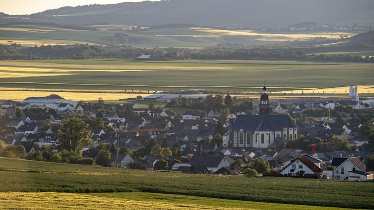 Eine ruhige Dorfansicht mit vielen Häusern und einer Kirche im Vordergrund. Im Hintergrund erstrecken sich sanfte Hügel und grüne Felder.