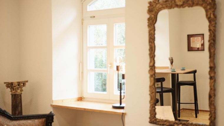 A bright room with a window and a mirror on the wall.
Next to the window, there is a table with chairs.