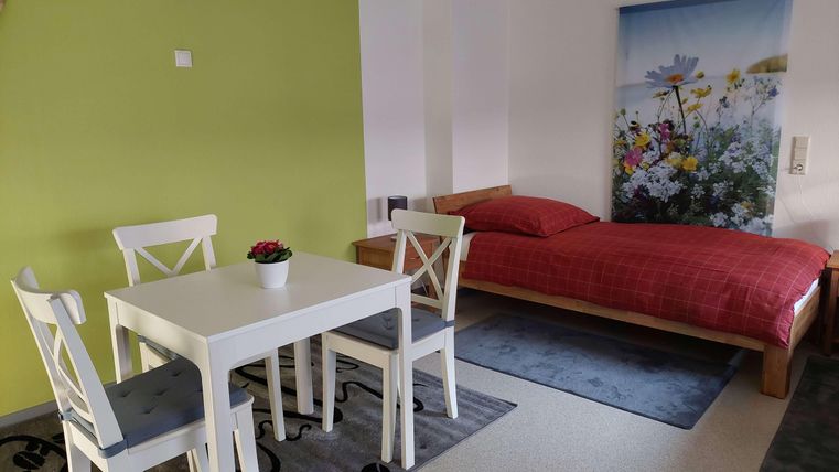 A cozy room with a red bed and a small dining table. On the wall, there is a colorful flower picture.
