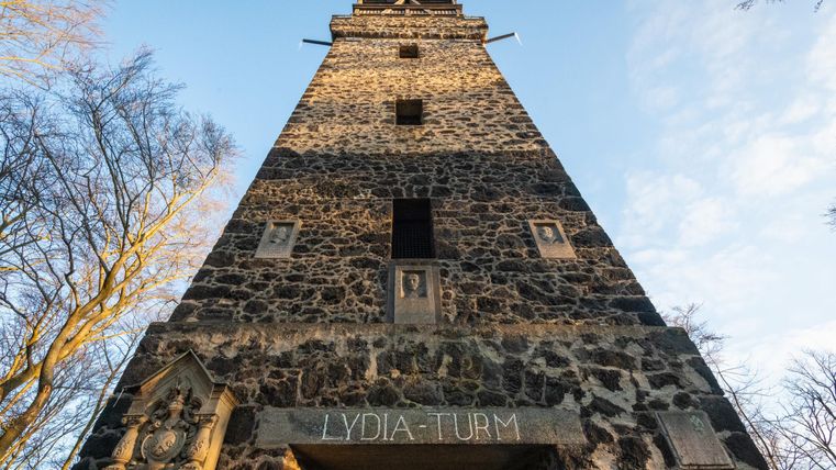 A tall stone tower with the inscription "LYDIA-TURM". Surrounded by trees and a clear sky.