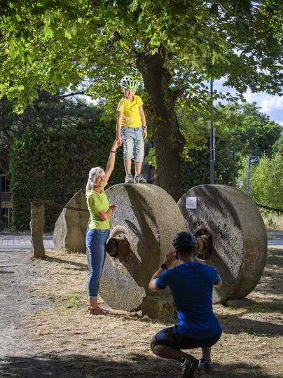 Ein Junge steht auf einem großen Steinrad und wird von einer Frau unterstützt. Im Hintergrund steht eine Person, die ein Foto macht.