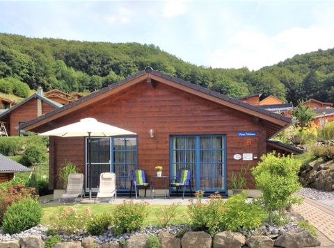 A cozy wooden house with a well-kept garden. In the foreground, there are lounge chairs and a sun umbrella.