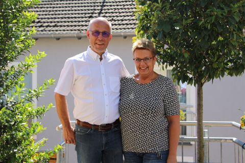 A friendly couple is standing and smiling in a garden. In the background, trees and a building can be seen.