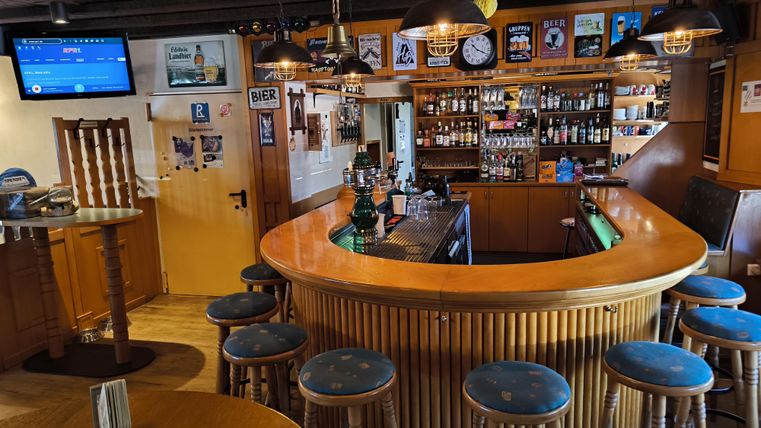 An inviting bar with a round counter and blue stools. In the background, numerous bottles and drinks are ready.