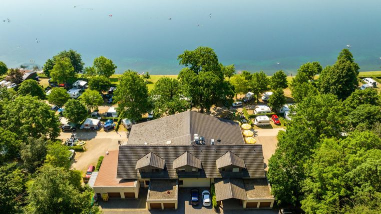 Ein malerisches Campingszenario am Ufer eines Sees, umgeben von Bäumen. Im Vordergrund ist ein Gebäude mit mehreren Dächern und parkenden Fahrzeugen zu sehen.