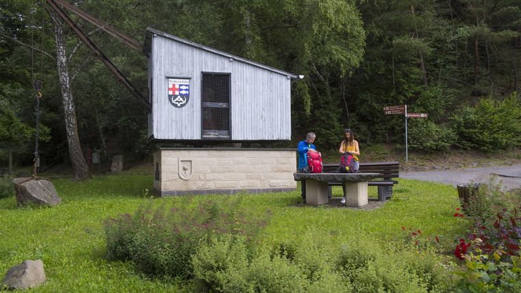 Ein Holzhaus auf einer Wiese, umgeben von Bäumen. Zwei Personen stehen neben einem Tisch und schauen in die Ferne.